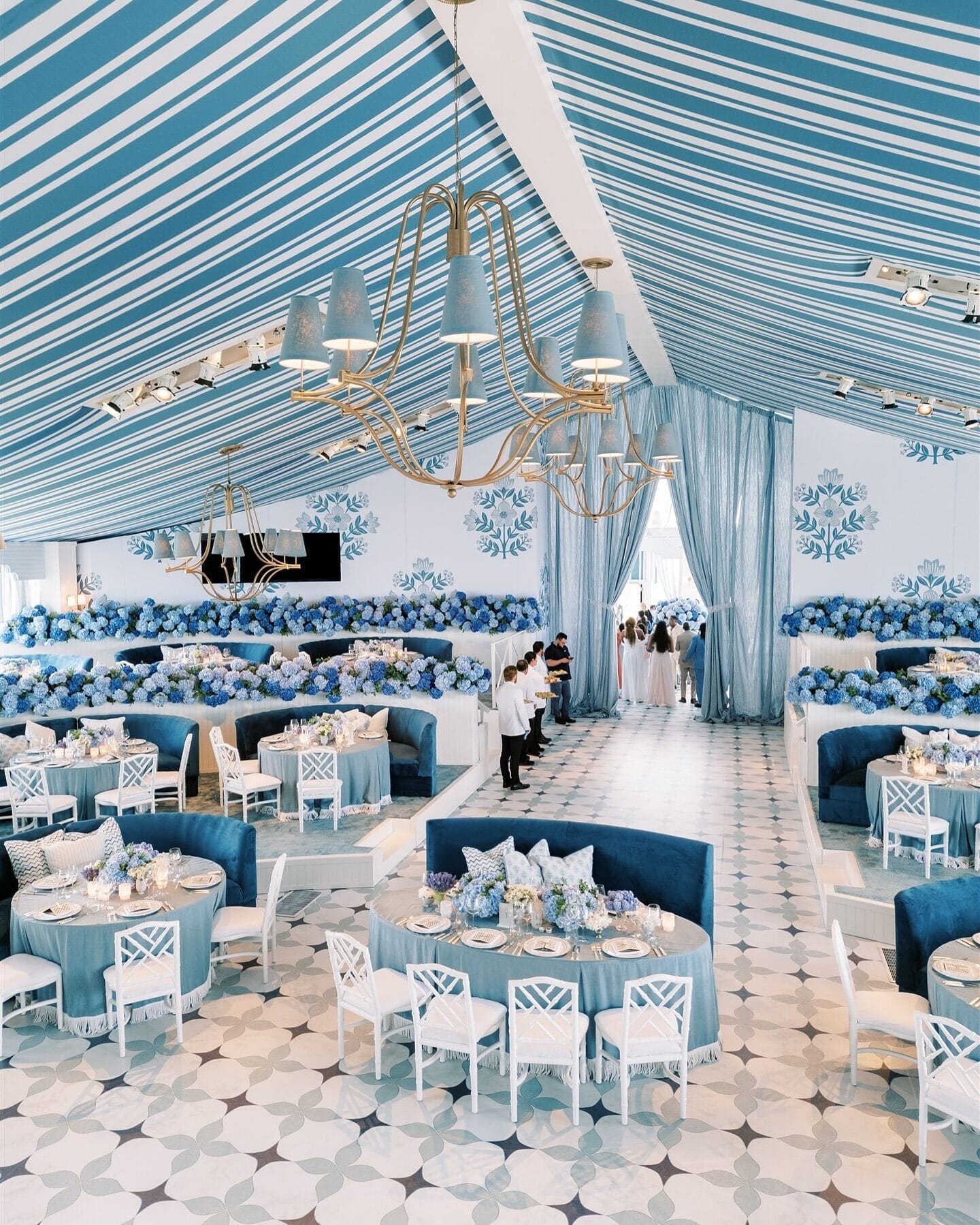 An opulent wedding reception space designed with blue and white tones, featuring custom ceiling drapery, elegant chandeliers, floral-patterned walls, and plush seating. The layout showcases a well-curated blend of sophistication and personalized design, reflecting the expertise of a luxury wedding planner. When choosing the perfect wedding planner for your luxury wedding, look for someone who can execute a cohesive vision, ensuring every detail aligns seamlessly with your style and grandeur.