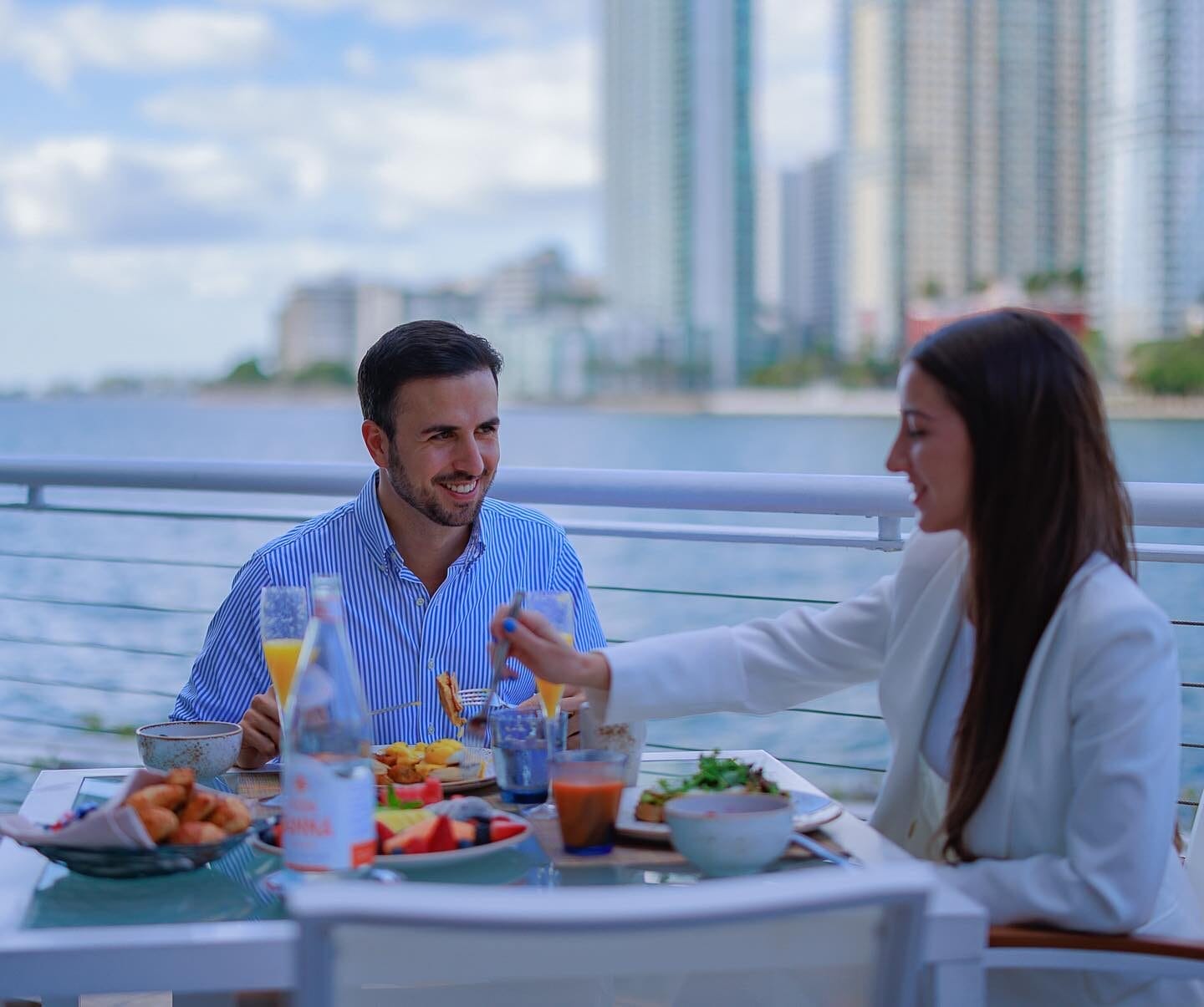 A couple enjoys a waterfront breakfast at Mandarin Oriental, Miami, with stunning views of Biscayne Bay and the city skyline. The hotel's serene setting and elegant outdoor dining create a luxurious and romantic atmosphere.