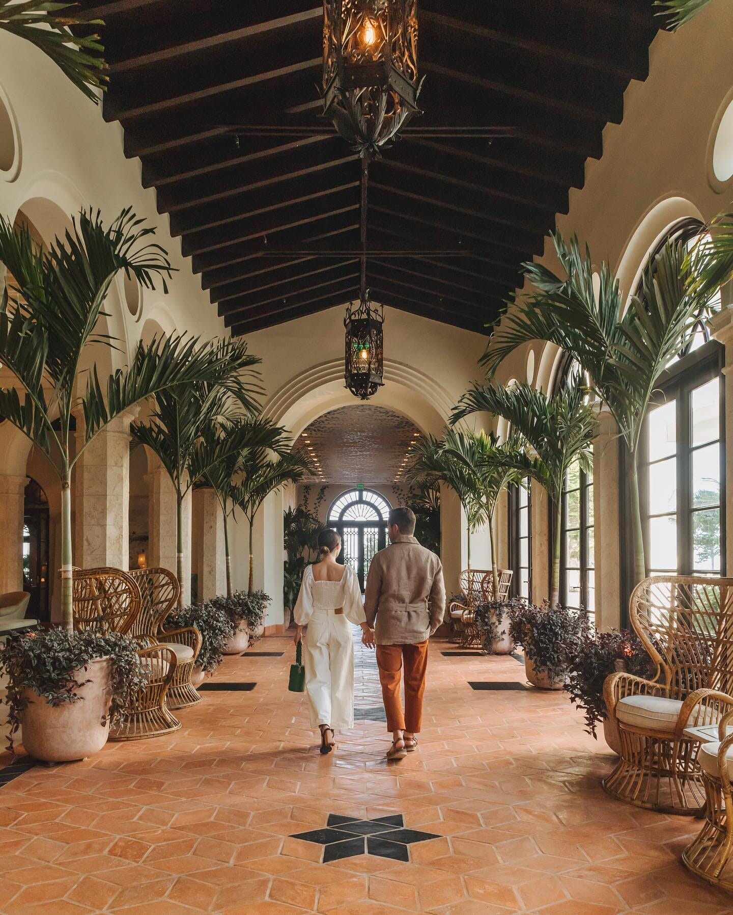A couple walks hand in hand through a beautifully designed corridor at Four Seasons Hotel at The Surf Club, surrounded by arched windows, lush greenery, and elegant lanterns. With its historic charm and refined luxury, the hotel provides a serene and sophisticated retreat for couples.